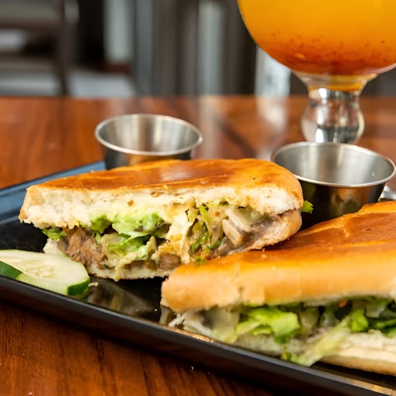 A Mexican torta sandwich, cut in half, with lettuce, meat, and sauce, served with cucumber slices.