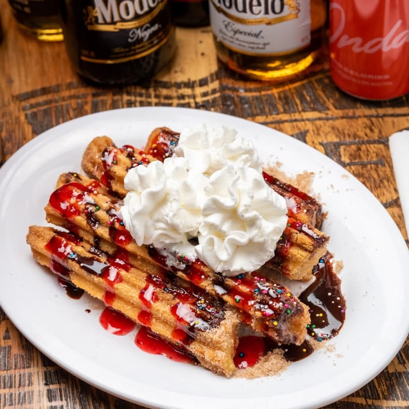 Churros with whipped cream, chocolate, strawberry syrup, and sprinkles on a white plate.