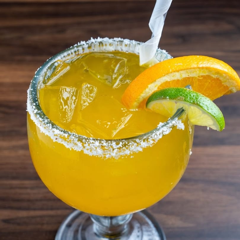 A vibrant orange margarita with ice, garnished with lime and orange slices, and a salted rim.